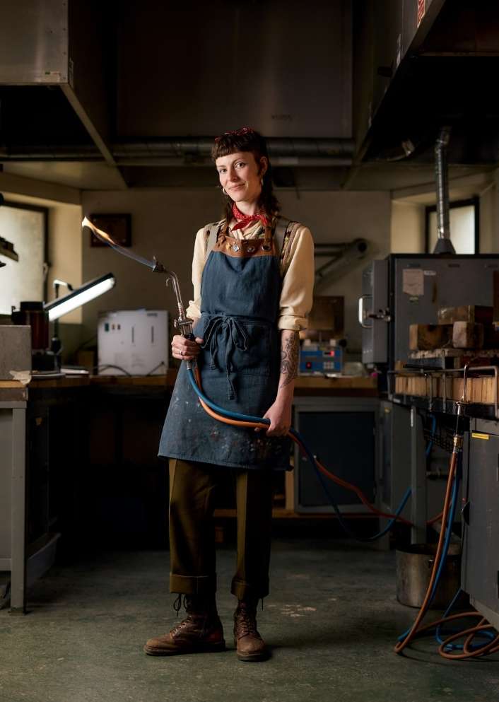 Full shot of a young adult woman holding a blowtorch, wearing a denim apron, and standing in an industrial workshop.