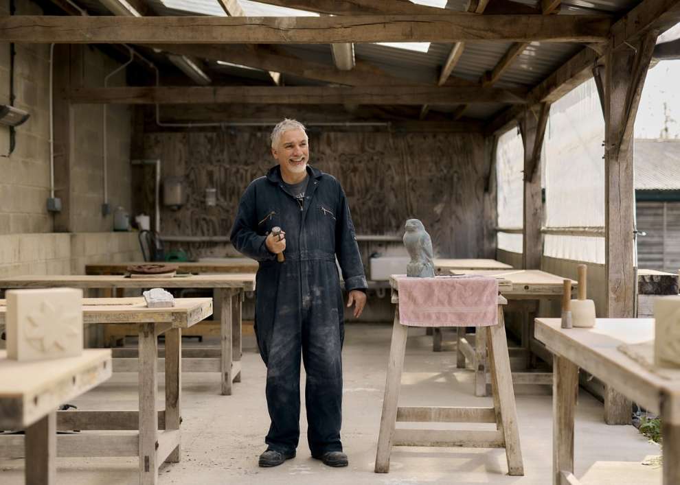 Smiling man in overalls stands in a workshop, holding a hammer. A small bird sculpture sits on a table next to him.