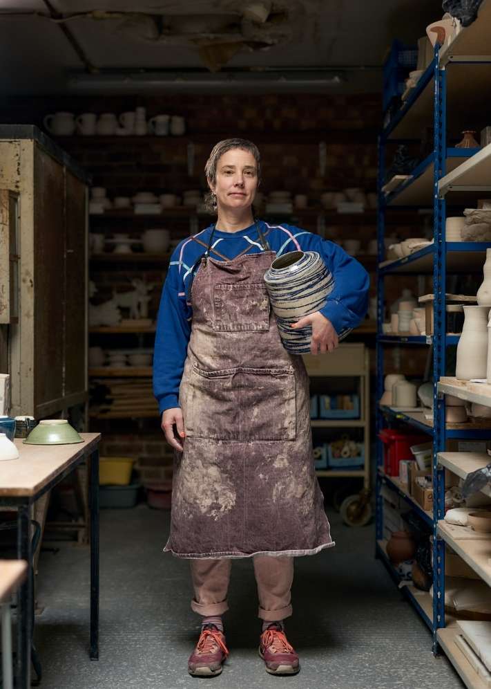 Adult woman in a workshop holding a vase, wearing a faded apron, and pink pants, surrounded by shelves with pottery and art supplies.