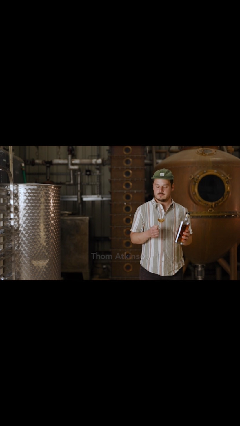 Man in patterned shirt stands in a distillery holding a glass and bottle, surrounded by stainless steel tanks and copper distilling equipment.