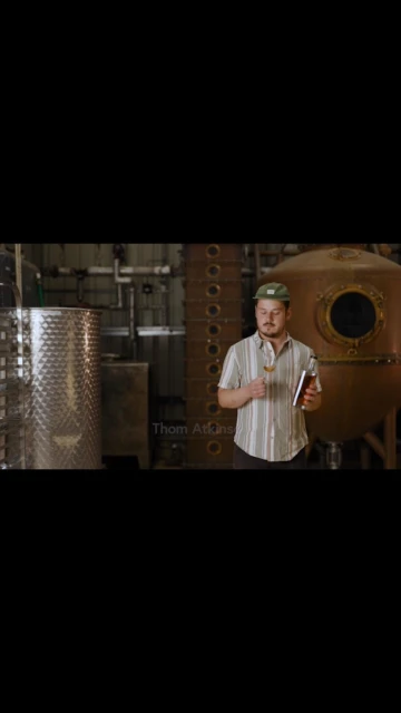 Man in patterned shirt stands in a distillery holding a glass and bottle, surrounded by stainless steel tanks and copper distilling equipment.