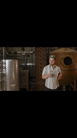 Man in patterned shirt stands in a distillery holding a glass and bottle, surrounded by stainless steel tanks and copper distilling equipment.