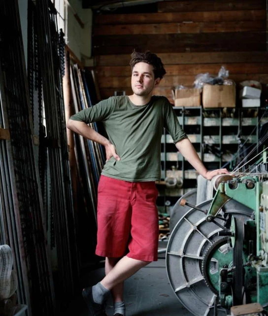 Male model in olive green shirt and red shorts, leaning against a large weaving machine in a workshop with natural window light.