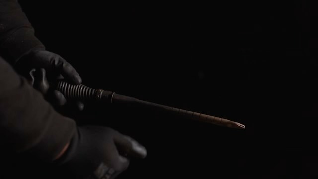 Gloved hands manipulating a cylindrical, dark brown object in dim lighting against a black background, creating a mysterious atmosphere.