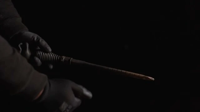 Gloved hands manipulating a cylindrical, dark brown object in dim lighting against a black background, creating a mysterious atmosphere.