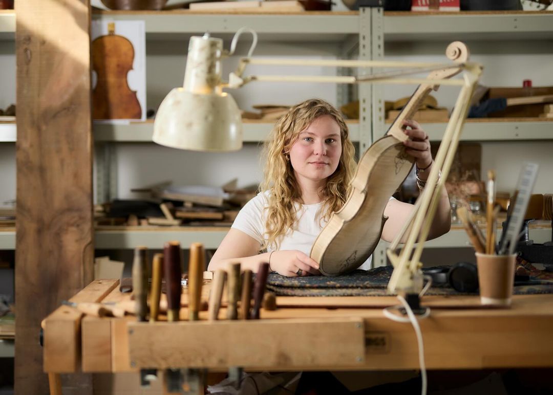 Woman crafting a violin in a workshop: Blonde woman holds unfinished violin body at a workbench with tools in a brightly-lit workshop.