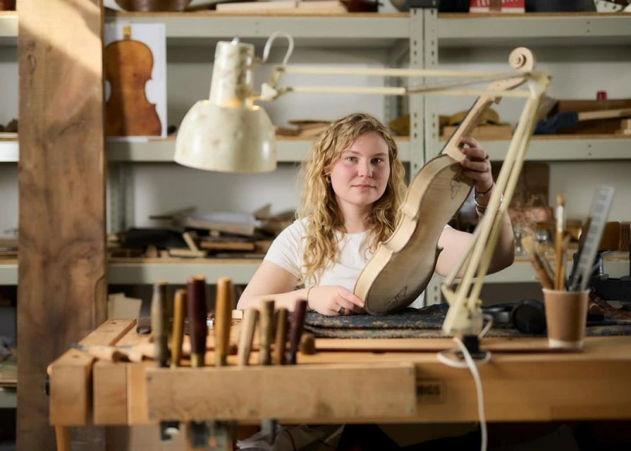 Woman crafting a violin in a workshop: Blonde woman holds unfinished violin body at a workbench with tools in a brightly-lit workshop.