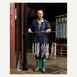 A man in a dark jumpsuit and green rubber boots holds a horizontal stick from which many silver spoons hang downward by wire in a factory.