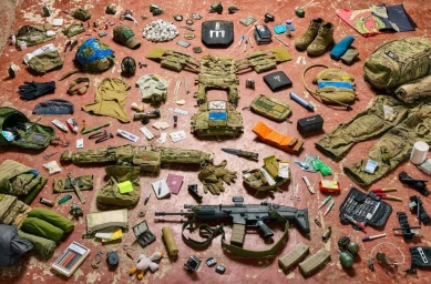 Overhead flat lay photograph showing military gear, assault rifle, tactical vest, helmet, and equipment arranged on a textured reddish floor.