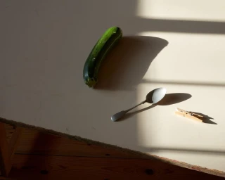 Green zucchini, silver spoon, and wooden clothespin on a pale table under slatted light.