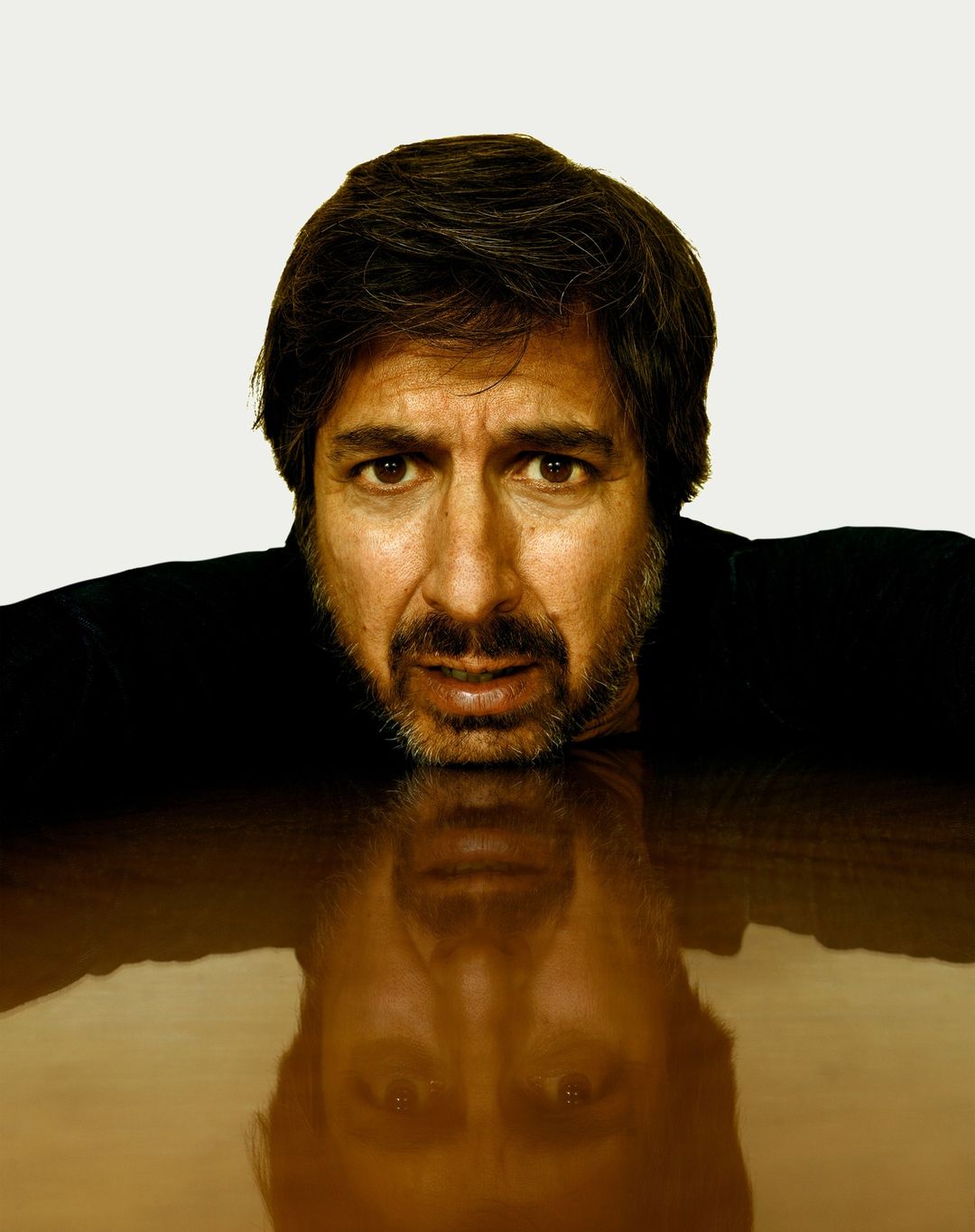 Headshot of a middle-aged man with dark hair and beard leaning on a brown surface, reflected below, set against a white background.
