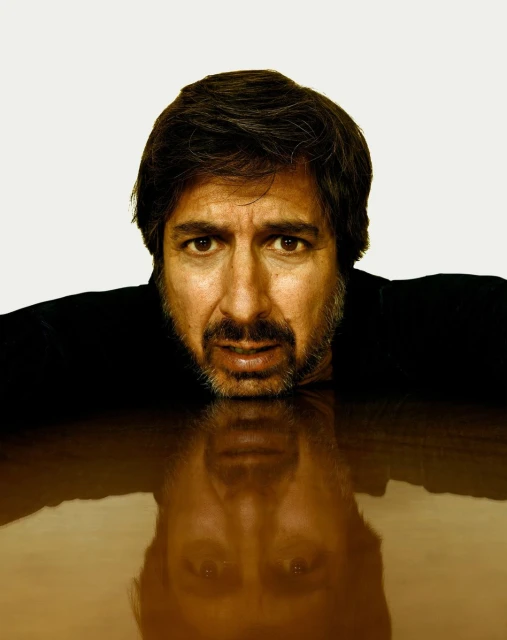 Headshot of a middle-aged man with dark hair and beard leaning on a brown surface, reflected below, set against a white background.