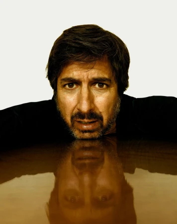 Headshot of a middle-aged man with dark hair and beard leaning on a brown surface, reflected below, set against a white background.