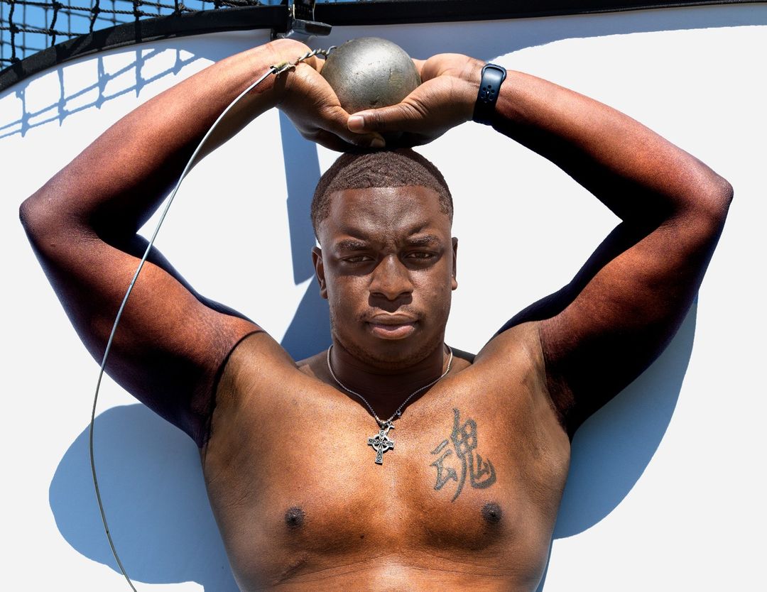 African-American male athlete in sunlight. He is holding a metal shot put above his head with both hands.