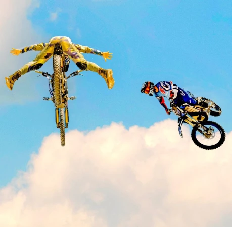 Two motocross riders in mid-air against a bright blue sky with light clouds, performing stunts with their bikes. One rider is upside down.