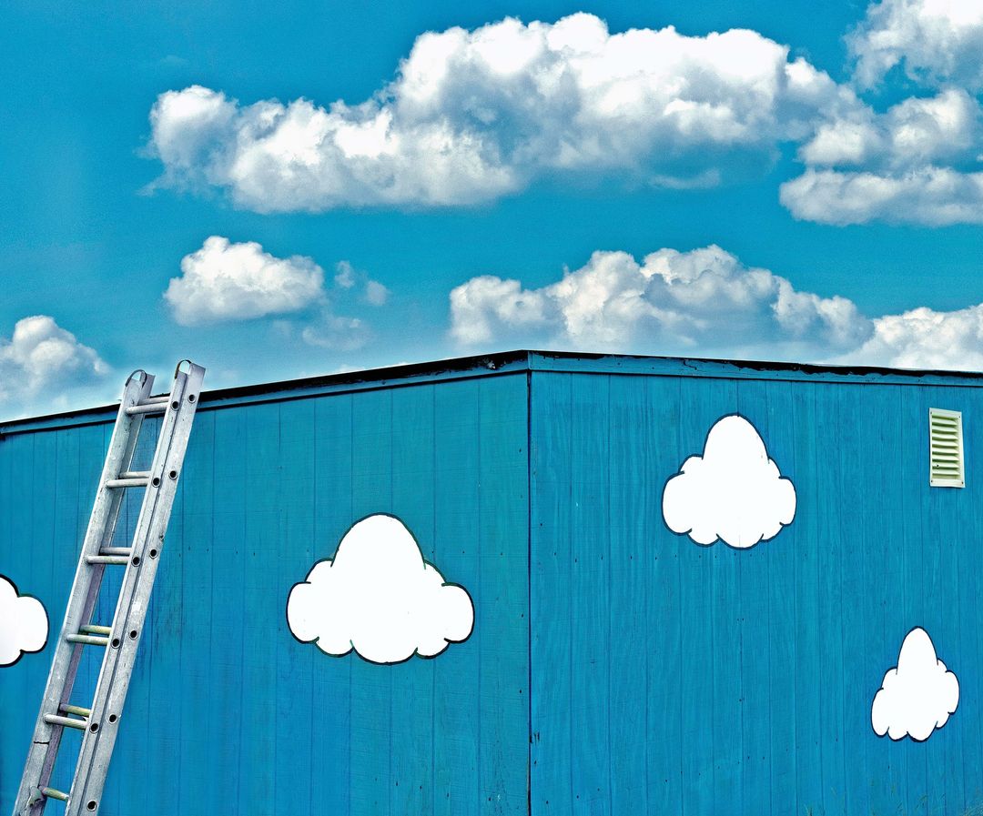 A blue building with white clouds painted on the side and a silver ladder leaning against the building under a blue sky with clouds.