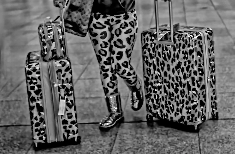 A person in leopard-print leggings walks with matching suitcases on a tile floor in this high-contrast black-and-white photograph.