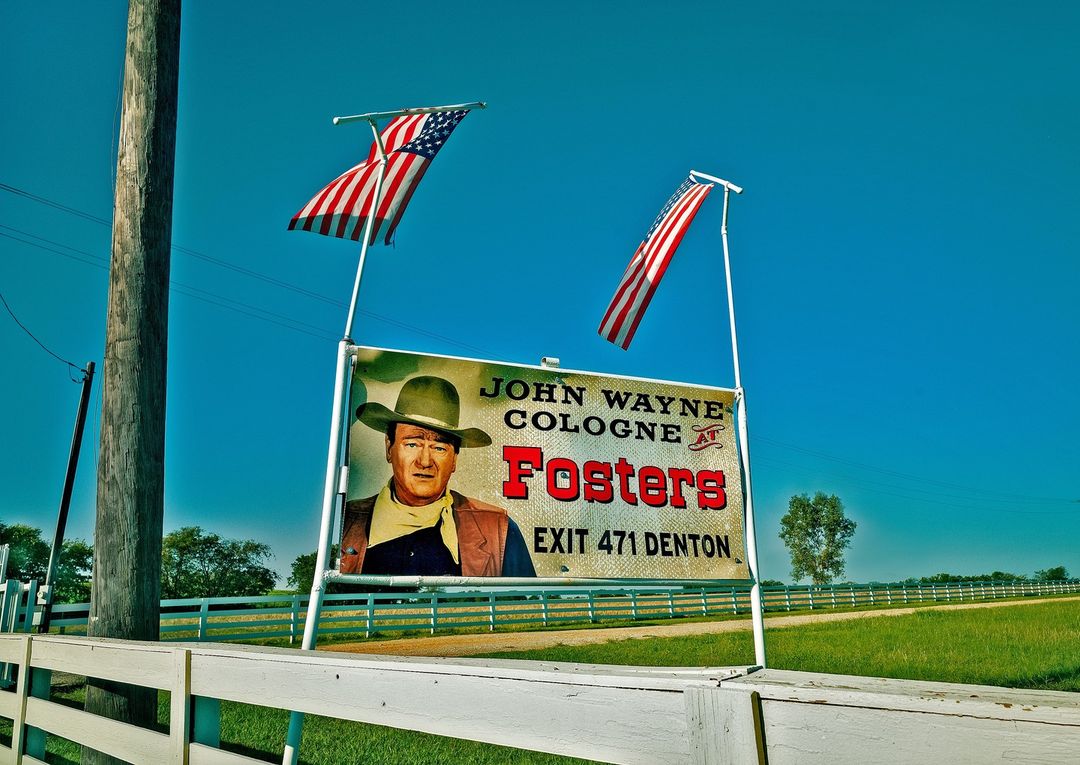 Vintage billboard of John Wayne cologne, American flags, white fence, and bright blue sky in the Texas countryside during daytime.