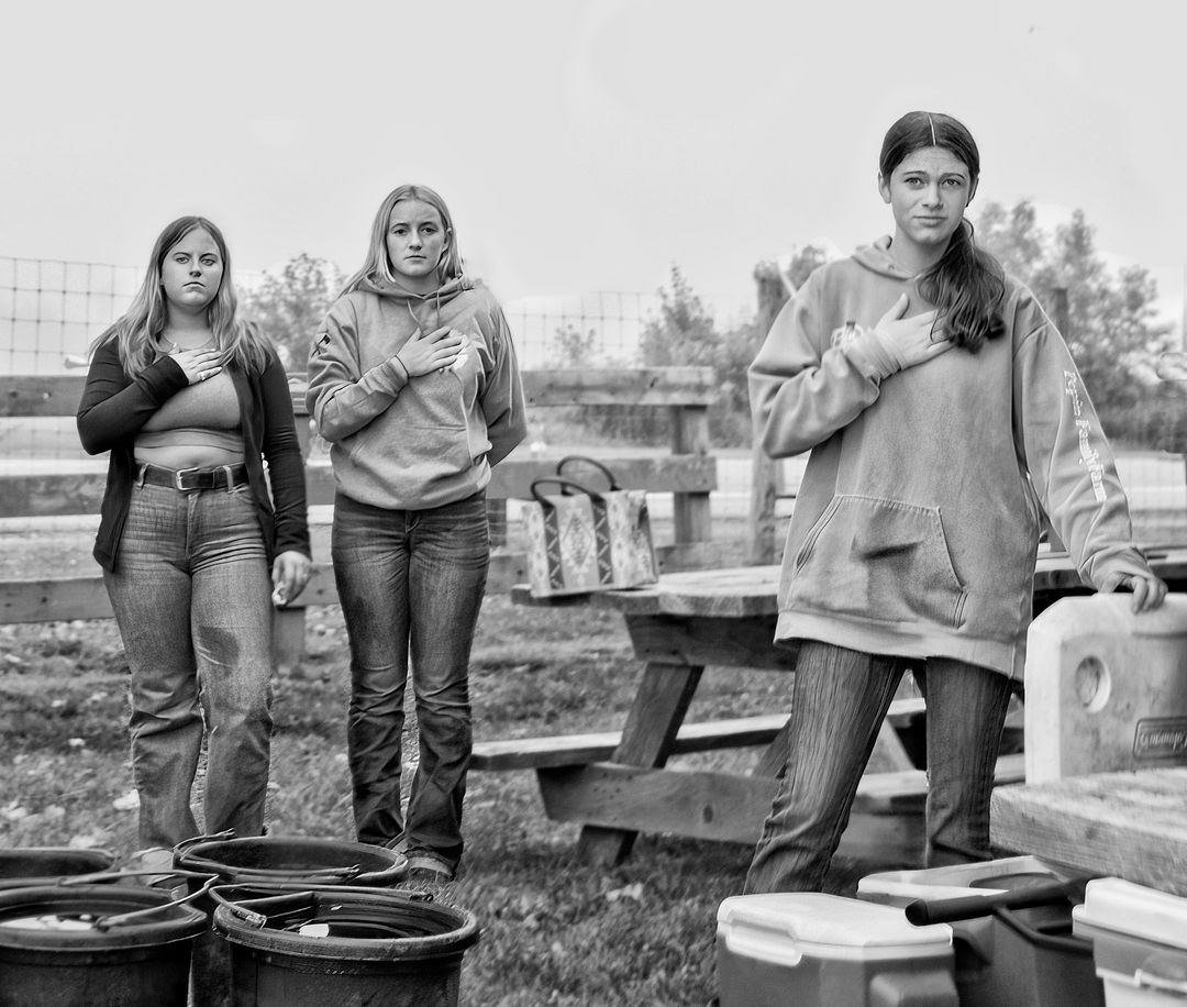 Three young women in jeans and hoodies stand outdoors with hands over their hearts, in a black and white photo on a cloudy day.