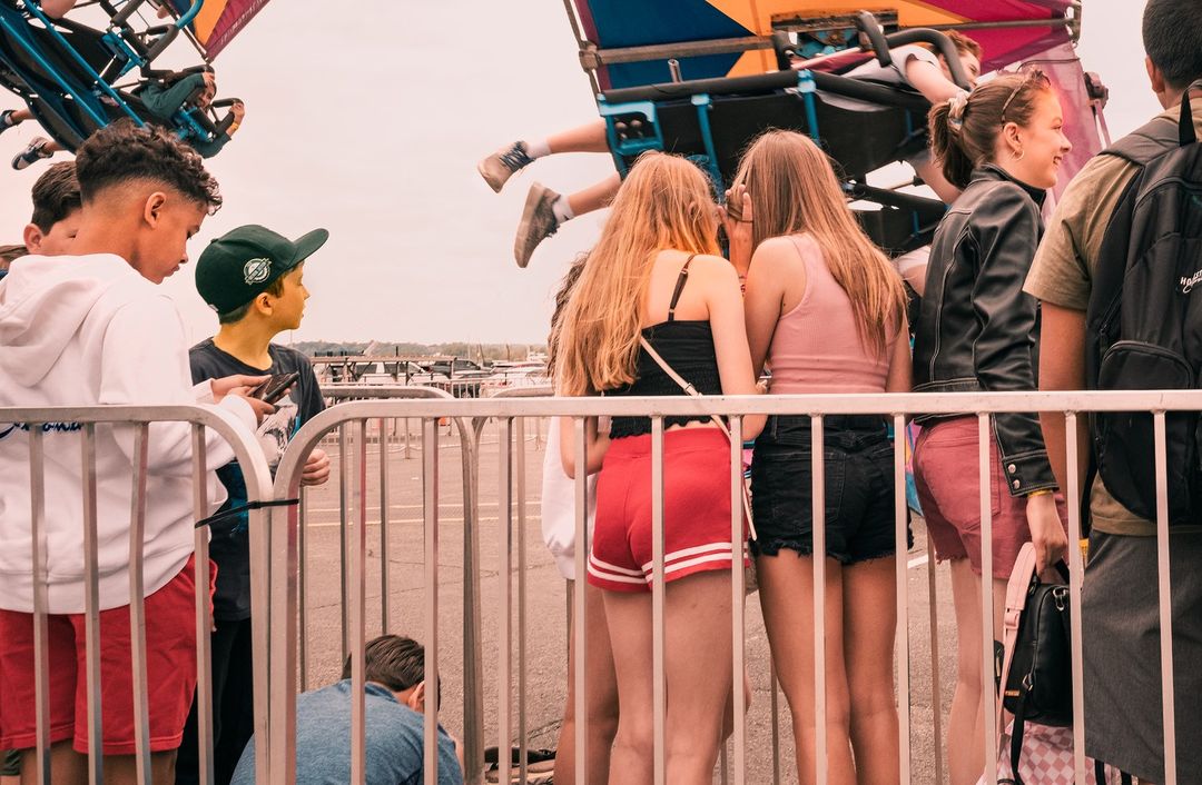 Crowd of young people in shorts and casual clothes stand by metal barrier at an outdoor fair, watching a carnival ride on a cloudy day.