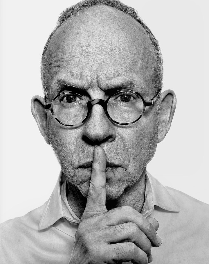 Portrait of a mature man with glasses holding a finger to his lips, making a 'shhh' gesture, set against a white background in black and white.