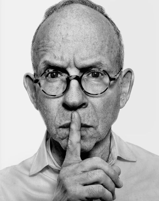 Portrait of a mature man with glasses holding a finger to his lips, making a 'shhh' gesture, set against a white background in black and white.