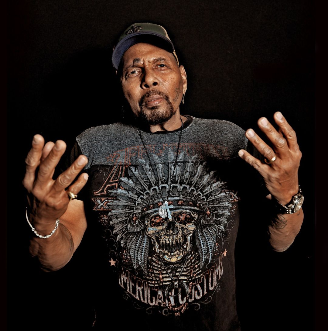 Portrait of an elderly man wearing a graphic t-shirt and baseball cap, holding both hands up against a black background.