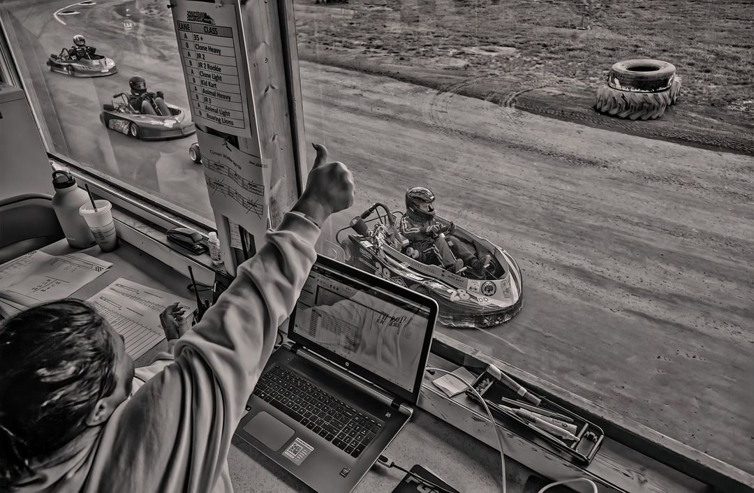 Monochrome shot of go-kart racers speeding on an outdoor track, viewed from a race official's booth with visible laptop and paperwork.