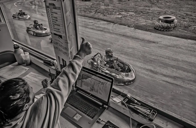 Monochrome shot of go-kart racers speeding on an outdoor track, viewed from a race official's booth with visible laptop and paperwork.