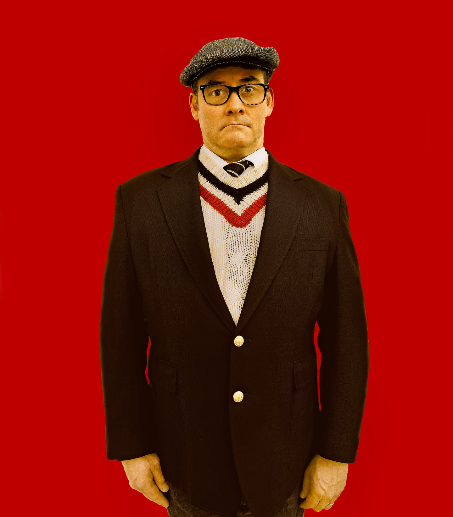 A man with glasses and a newsboy cap wears a black blazer over a white sweater with a red and blue V-neck against a red background.