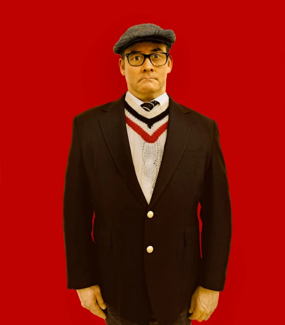 A man with glasses and a newsboy cap wears a black blazer over a white sweater with a red and blue V-neck against a red background.