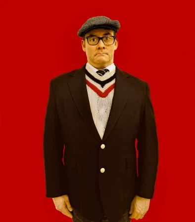 A man with glasses and a newsboy cap wears a black blazer over a white sweater with a red and blue V-neck against a red background.