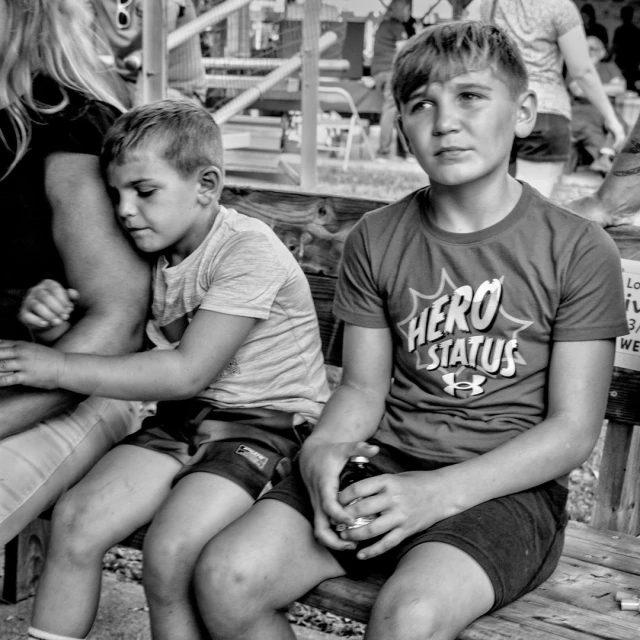 Two young boys sit on a bench in a black and white outdoor photograph. One boy rests on an adult’s arm while the other holds a bottle.