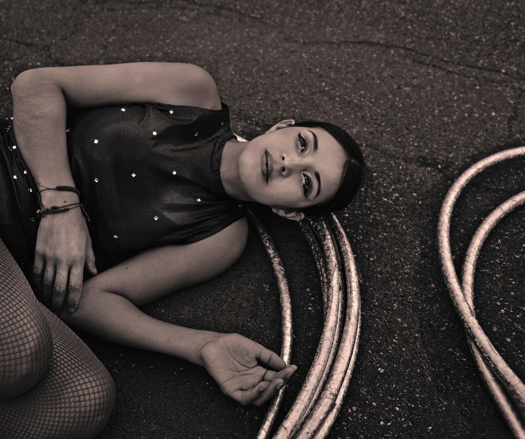 Woman in shimmery bodysuit lays on concrete next to hula hoops, with a sepia tone for a vintage editorial-style image.