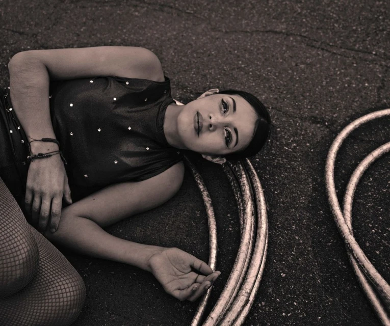 Woman in shimmery bodysuit lays on concrete next to hula hoops, with a sepia tone for a vintage editorial-style image.