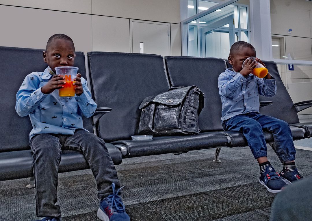 Two young boys, dressed in button-down shirts and dark jeans, sit side-by-side on chairs at an airport while holding and drinking orange juice.