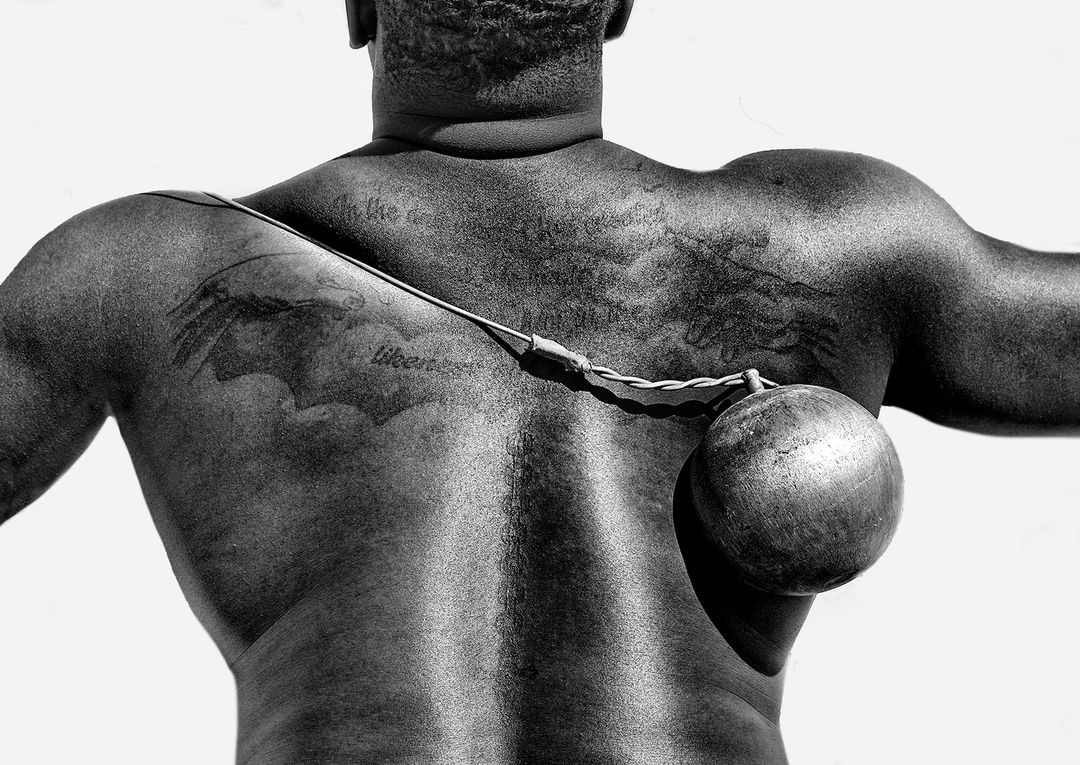 Back view of adult male athlete with tattoos holding a metal hammer ball with cable and handle, set against a bright, neutral backdrop.