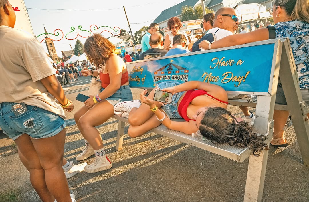 Several people are at an outdoor fair or festival with a young woman laying across a bench looking at her cell phone screen.