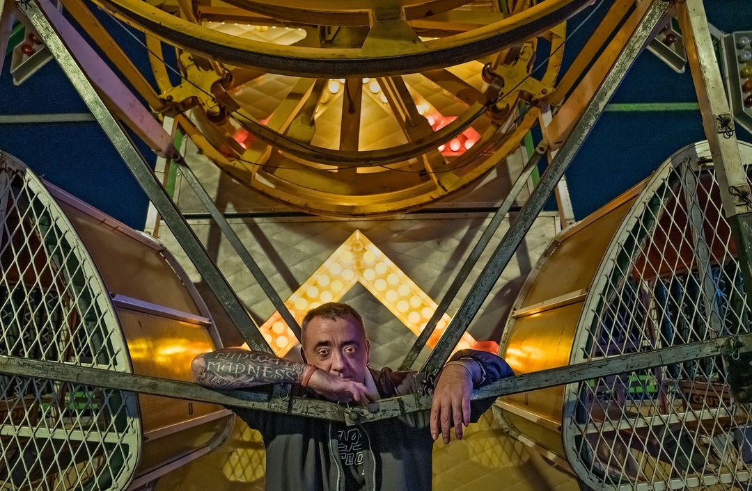 Man poses near a brightly lit Ferris wheel at night, showcasing tattoos and casual wear in an amusement park setting.