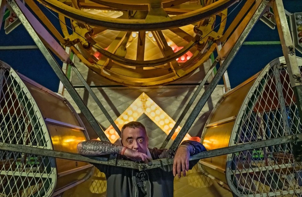 Man poses near a brightly lit Ferris wheel at night, showcasing tattoos and casual wear in an amusement park setting.