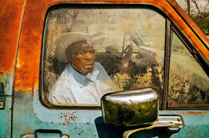 Older African-American man with a cowboy hat sits inside of an old, rusted pick-up truck with a reflective side mirror visible.