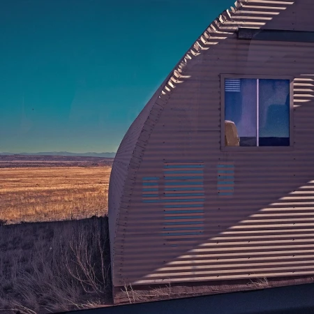 Exterior view of a corrugated metal structure with a window, set against a backdrop of clear blue sky and arid landscape. Desert scene.