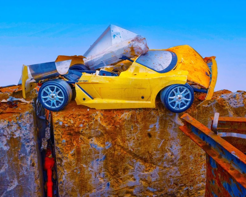 Damaged yellow toy car sits on a cracked orange surface with a blue sky background, showcasing a scene of destruction and weathering.