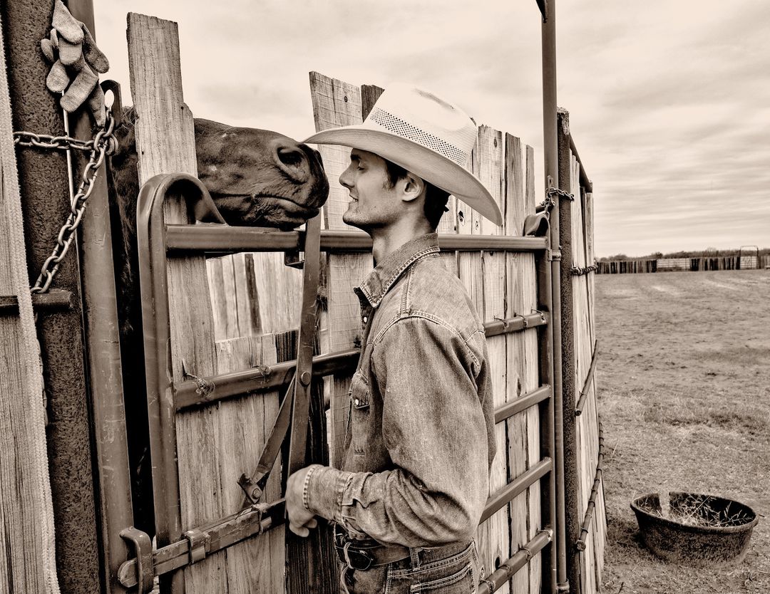 A sepia-toned image shows a cowboy in a hat and denim shirt, facing a brown horse in a wooden pen on a ranch under a cloudy sky.