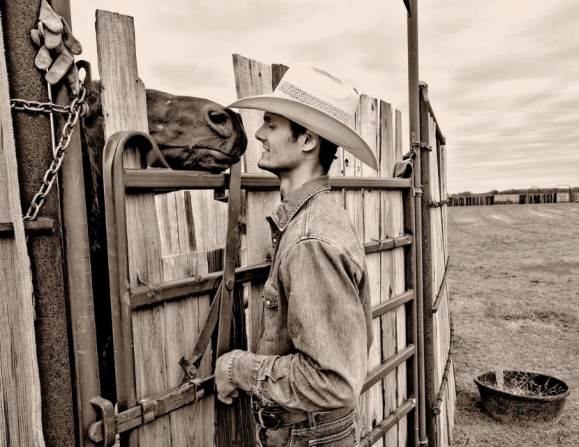 A sepia-toned image shows a cowboy in a hat and denim shirt, facing a brown horse in a wooden pen on a ranch under a cloudy sky.