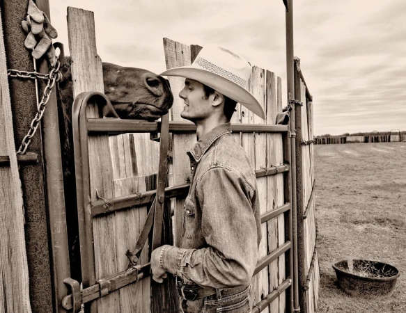 A sepia-toned image shows a cowboy in a hat and denim shirt, facing a brown horse in a wooden pen on a ranch under a cloudy sky.