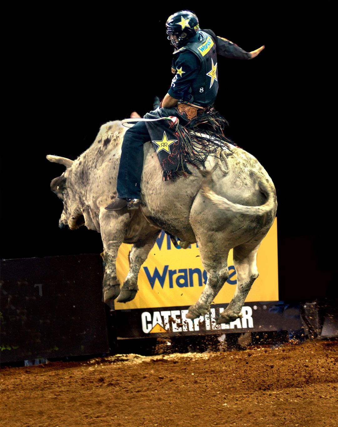 Bull rider in mid-air on a bucking bull, wearing protective gear and a star-studded vest, with arena signage visible in the background.