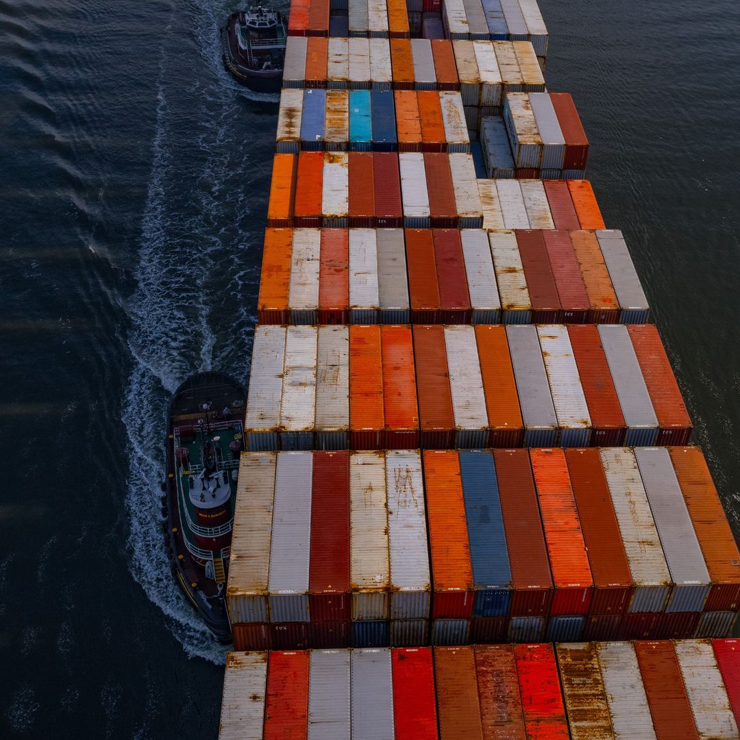 High-angle view of a large cargo ship with rows of colorful shipping containers, accompanied by two tugboats in the dark water.
