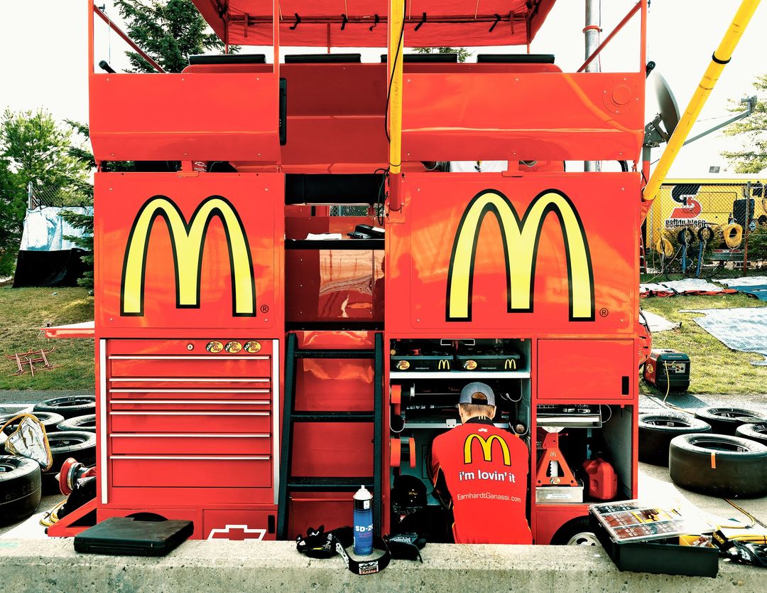 Race car pit stop with a mechanic working on a red car with McDonald's logo and tires stacked nearby outdoors on a sunny day.