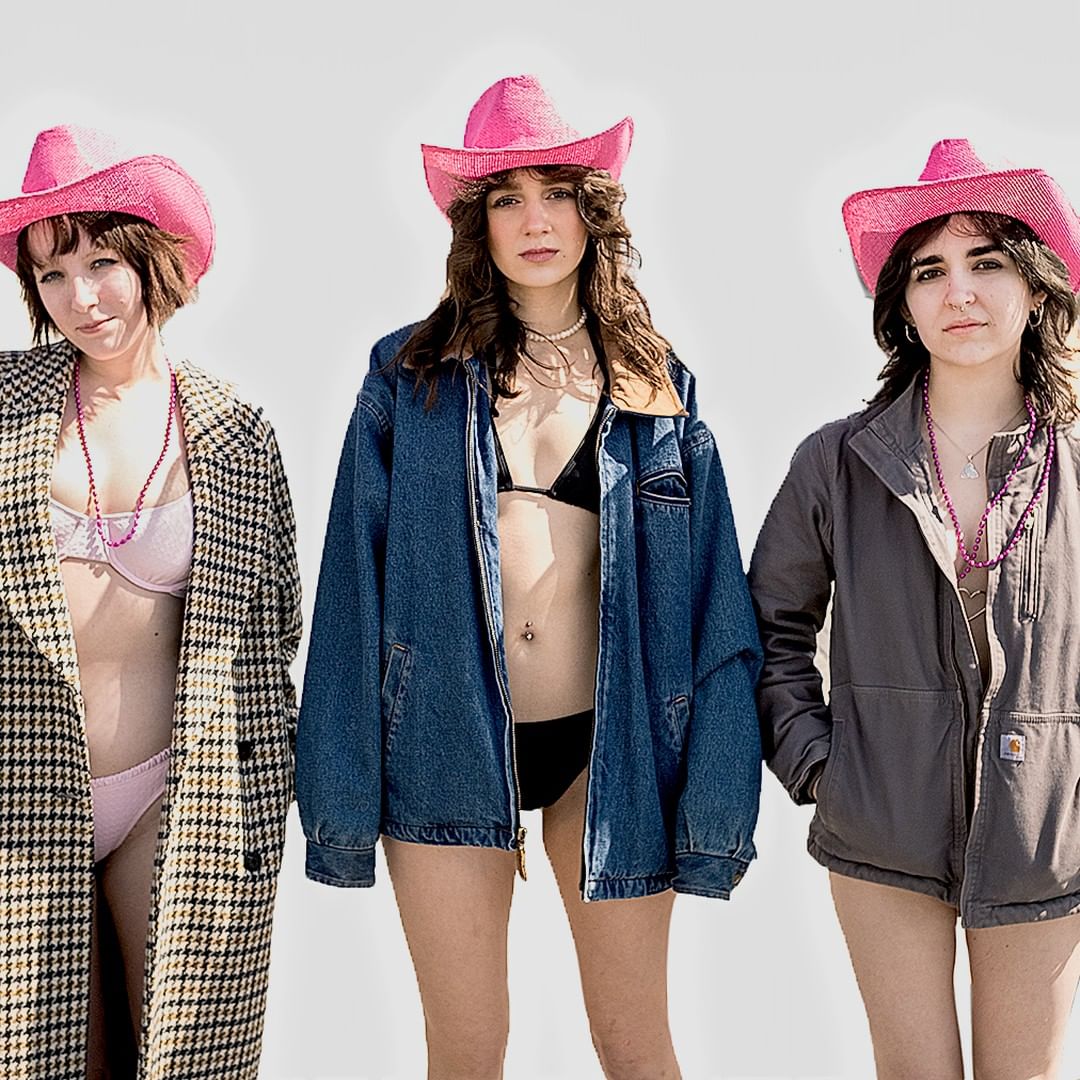 Three women in swimsuits and pink cowboy hats stand against a white backdrop, two wearing jackets, showcasing a blend of fashion styles.